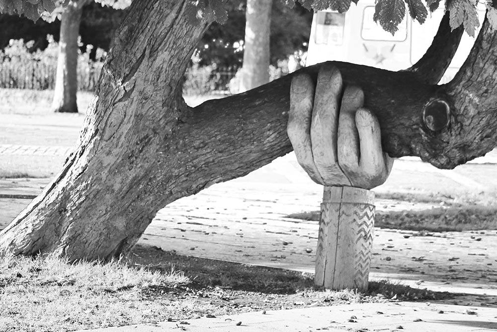 Eine aus Holz gefertigte große Hand stützt einen quer verlaufenden dicken Ast eines schräg wachsenden Baums.