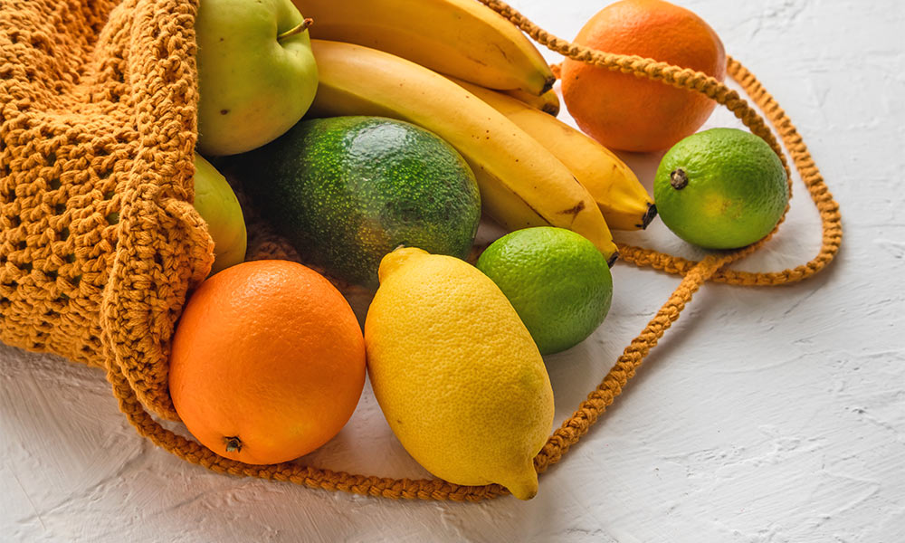 ein Einkaufsnetz mit Orangen, Bananen, Äpfeln, Zitronen, Limetten und einer Avocado.